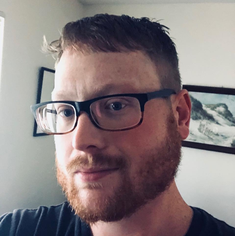 A close-up of a man with short brown hair, glasses, and a beard, smiling slightly in a room with artwork on the wall.