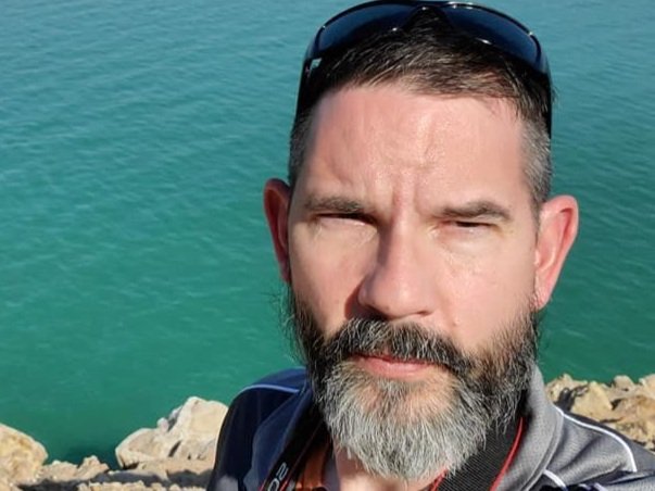 A man with a beard and sunglasses on his head, taking a selfie near a body of water with rocks in the background.