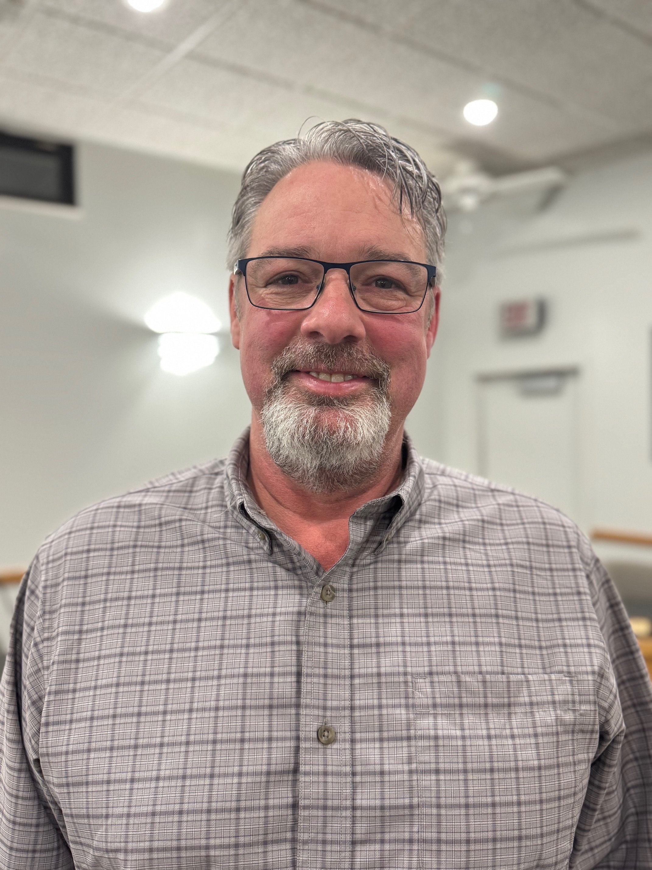 A middle-aged man with gray hair, glasses, and a beard, smiling in an indoor setting with white walls and ceiling lights.