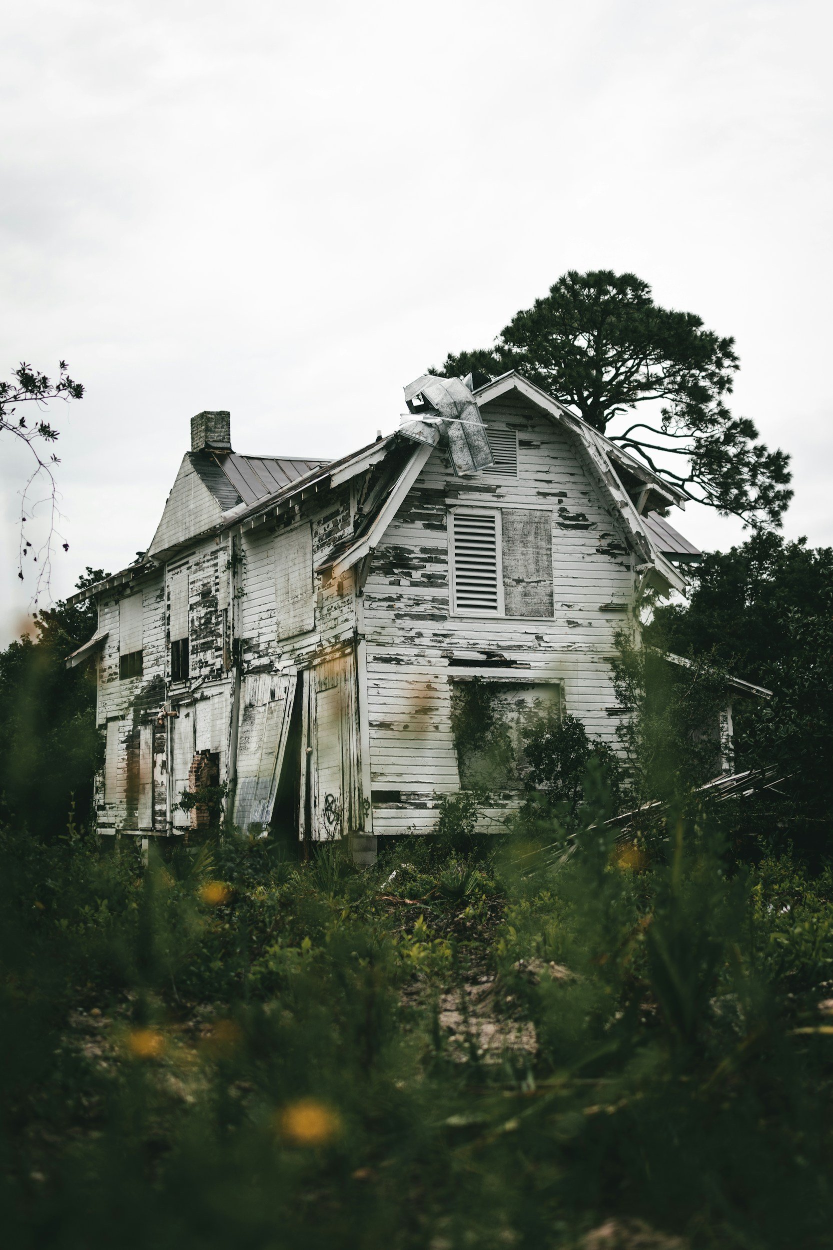 A dilapidated, abandoned house with peeling white paint and broken windows, surrounded by overgrown vegetation and a cloudy sky.