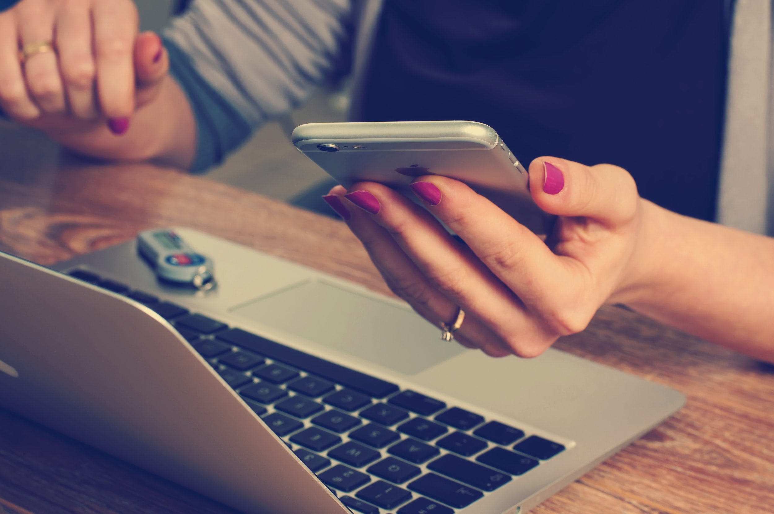 A woman with pink nail polish holding a smartphone while sitting at a wooden table with a laptop, car key, and a remote control visible on the table.