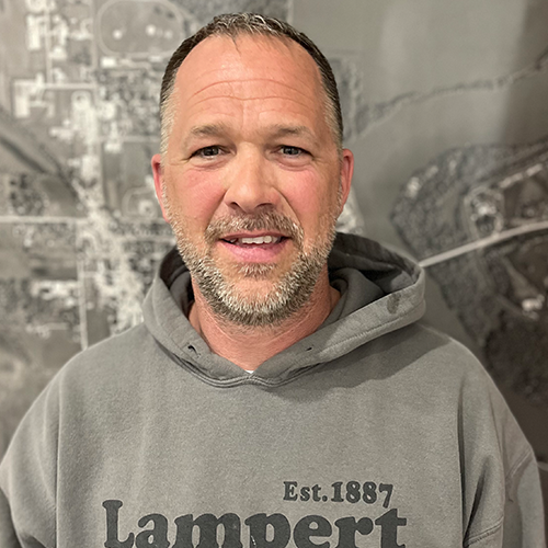 A smiling middle-aged man with a beard and short hair, wearing a gray hoodie with the word "Lampert" and "Est. 1887" printed on it, standing in front of a large black-and-white map or aerial photo of a city.