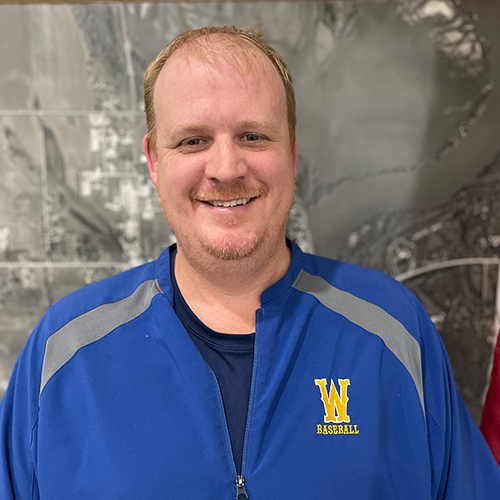 A smiling man with short light hair and a goatee, wearing a blue baseball jacket with a yellow 'W' and 'Baseball' embroidered on it, standing in front of a gray background.